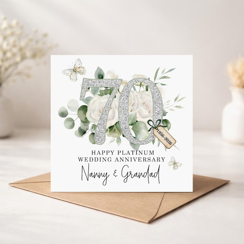 May include: A white card with a silver glitter "70" and the text "Happy Platinum Wedding Anniversary Nanny & Grandad". The card features white roses, greenery, and butterflies. A small tag reads "1956-2026". The card is on a brown envelope.