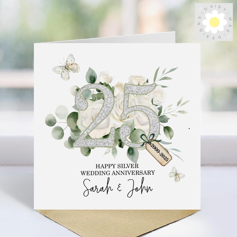 May include: A white card with a silver glitter number 25 surrounded by white roses and green leaves. The card says "Happy Silver Wedding Anniversary Sarah & John" and has a small tag that says "2000-2025".