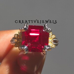 Op de afbeelding: Close-up van een ring met een grote, vierkant geslepen, felrode edelsteen. De ring heeft een zilveren band met gouden en zilveren decoratieve accenten. De tekst "CREATIVEJEWELS" is zichtbaar.