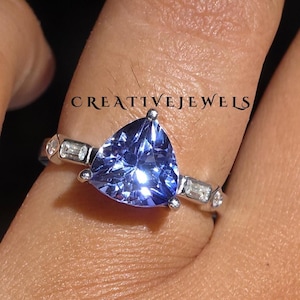 Op de afbeelding: Een close-up van een zilveren ring met een grote, driehoekige, lichtblauwe edelsteen. De ring heeft kleinere, rechthoekige stenen langs de band. De tekst "CREATIVEJEWELS" is zichtbaar.