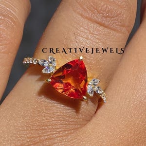 May include: A gold ring featuring a triangular, fiery orange gemstone as the centerpiece. The ring is adorned with smaller, clear and blue accent stones, and the band is embellished with additional small stones. The text "CREATIVEJEWELS" is visible.