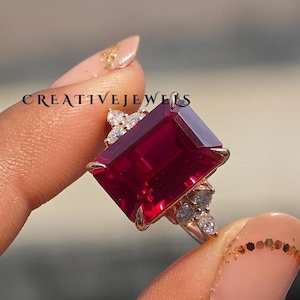 Puede incluir: Un anillo dorado con una gran gema rectangular de color rojo intenso. Gemas más pequeñas y claras acentúan la piedra principal y la banda del anillo. El texto "CREATIVEJEWELS" es visible.