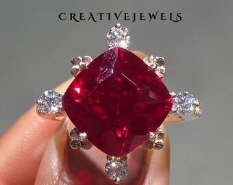 May include: A large, square-cut, deep red gemstone ring with smaller, clear accent stones. The ring is set in a silver or white gold band. The text "CREATIVEJEWELS" is above the ring.