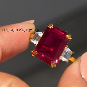 Puede incluir: Un anillo de oro con una gran gema rectangular de color rojo intenso como pieza central, flanqueada por dos gemas claras rectangulares más pequeñas. El anillo está sostenido por los dedos de una persona. El texto "CREATIVEJEWELS" es visible.