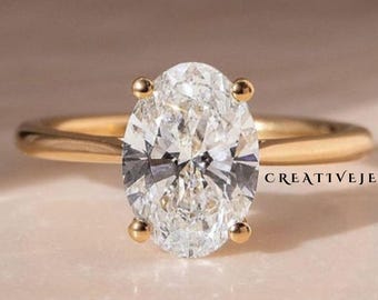 May include: A gold band ring with a large, oval-cut diamond. The diamond is clear and sparkling, set with four prongs. The ring is on a light pink surface. The text "CREATIVEJEWELS" is visible on the right side of the ring.