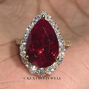 Könnte beinhalten: Ein goldener Ring mit einem großen, birnenförmigen, tiefroten Edelstein, umgeben von einem Kranz aus kleineren, klaren Edelsteinen. Der Ring hat ein goldenes Band. Der Text "CREATIVEJEWELS" ist sichtbar.