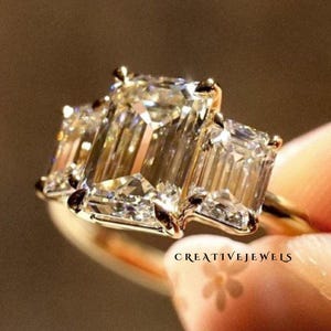 May include: A gold engagement ring with three emerald-cut diamonds. The central diamond is larger than the two flanking stones. The ring is held up against a blurred background, with the diamonds catching the light.