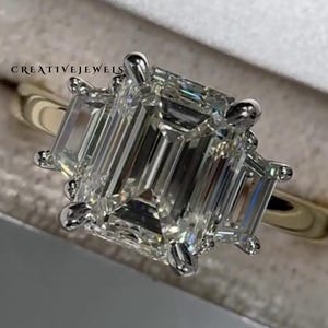 May include: A gold ring featuring a large, rectangular emerald-cut diamond, with two smaller emerald-cut diamonds on either side. The ring has a platinum or white gold setting. The brand name "CREATIVEJEWELS" is visible.
