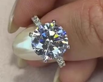 Pode incluir: Um anel de prata com uma grande pedra de diamante redonda, clara e brilhante no centro e diamantes menores ao redor da banda.