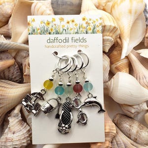 May include: A set of handcrafted stitch markers with beach-themed charms, including an umbrella, bucket, flip-flop, dolphin, and turtle. Each marker has a colorful bead. The card reads "daffodil fields handcrafted pretty things."