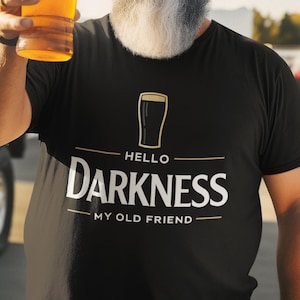 May include: Black t-shirt with the text "HELLO DARKNESS MY OLD FRIEND" in white and gold. The shirt features a beer glass graphic. A person is holding a glass of beer.
