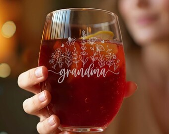 May include: A stemless wine glass filled with a red beverage and a lemon slice. The glass is etched with the word "grandma" in cursive, along with a floral design. The glass is held by a person.