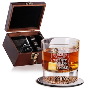 May include: A whiskey glass with the text "Retired 2025 Not My Problem Anymore" sits on a coaster. A wooden box with a glass and metal ice cubes is open in the background. The glass is filled with amber liquid.