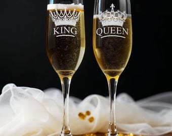 May include: Two champagne flutes with gold crowns and the words "KING" and "QUEEN" etched on them. The flutes are filled with champagne and are sitting on a table with gold confetti.