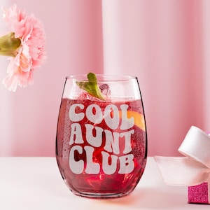 May include: A stemless wine glass filled with a red beverage, ice, and garnishes. The glass has the words "COOL AUNT CLUB" etched in white. A pink flower and a pink gift box with a white ribbon are in the background.