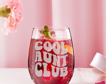 May include: A stemless wine glass filled with a red beverage, ice, and garnishes. The glass has the words "COOL AUNT CLUB" etched in white. A pink flower and a pink gift box with a white ribbon are in the background.