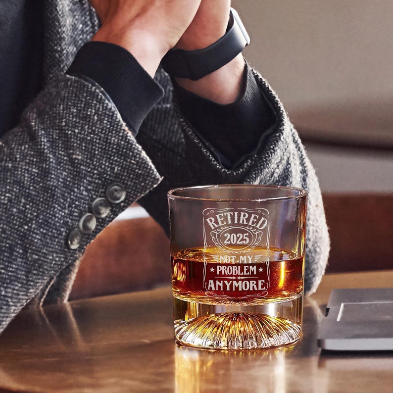 May include: A clear glass tumbler with a decorative base, filled with amber liquid. The glass is etched with the words "Retired 2025" and "Not My Problem Anymore". The glass is on a wooden surface.