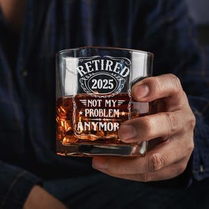 May include: A square whiskey glass filled with amber liquid and ice. The glass has white text that reads "RETIRED 2025" above "NOT MY PROBLEM ANYMOR". The glass is held by a person wearing a dark blue shirt.