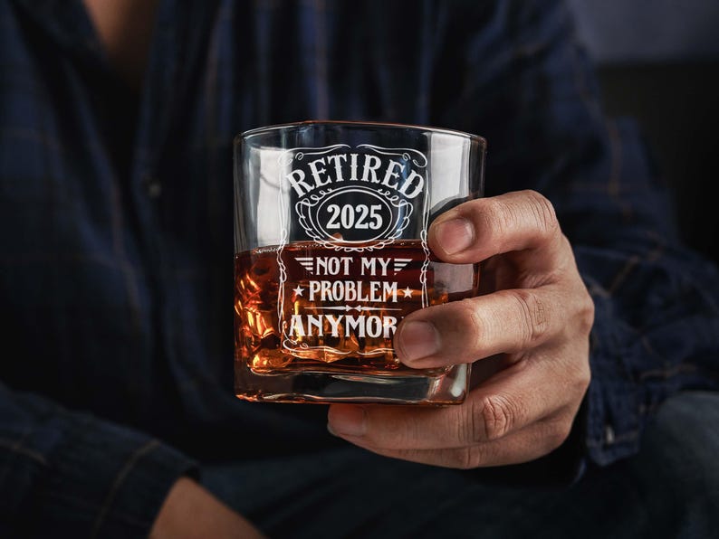 May include: A square whiskey glass filled with amber liquid and ice. The glass has white text that reads "RETIRED 2025" above "NOT MY PROBLEM ANYMOR". The glass is held by a person wearing a dark blue shirt.