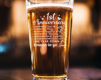 May include: A clear glass beer stein with a white inscription that reads "1st Anniversary 12 months of love, 52 weeks of bliss, 365 days of loyalty, 8760 hours of blessing, 525600 minutes of laughter, 31536000 seconds of friendship, One year down, Forever to go."