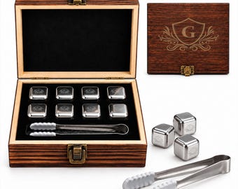 May include: A set of eight stainless steel whiskey stones with a letter G engraving, housed in a dark brown wooden box with a brass latch. The set includes a pair of metal tongs with white silicone tips, and the box lid features a matching G crest.