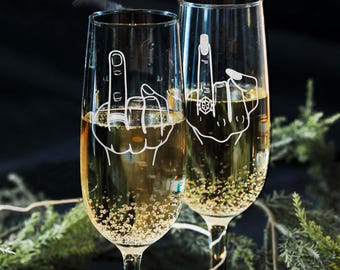 May include: Two champagne flutes with a humorous design etched on the glass. One flute features a middle finger with a ring on it, while the other flute features a hand with a ring on the index finger.