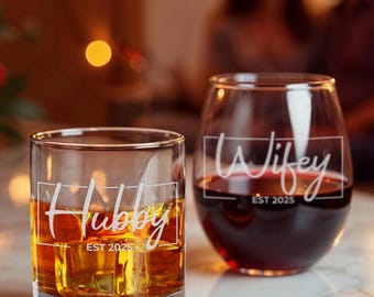 May include: Two clear glass tumblers with etched text. The tumbler on the left says "Hubby Est 2025" and is filled with amber liquid. The tumbler on the right says "Wifey Est 2025" and is filled with red wine.