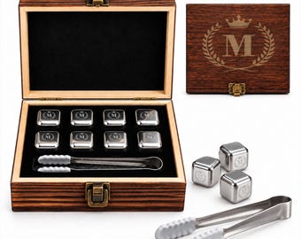 May include: A set of eight square stainless steel whiskey stones with a letter M and crown engraving, housed in a dark wood box with a metal latch. The set includes a pair of metal tongs with white silicone tips for handling the cubes.