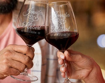 May include: Two clear glass wine glasses filled with red wine, each etched with the names Jennifer and Robert, two heart icons, and the date 25.11.2024. The glasses feature a tapered bowl, a slender stem, and a circular base for stability.