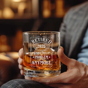 May include: A clear whiskey glass with the words "Retired 2025 Not My Problem Anymore" etched in white. The glass is filled with amber liquid and ice cubes. The glass is held by a person.