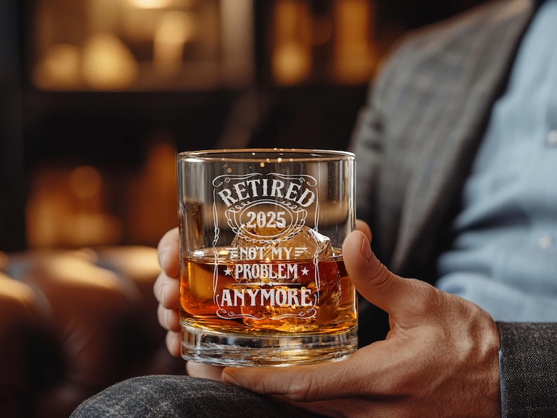 May include: A clear whiskey glass with the words "Retired 2025 Not My Problem Anymore" etched in white. The glass is filled with amber liquid and ice cubes. The glass is held by a person.
