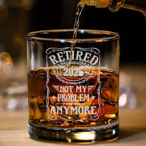 May include: A clear rocks glass filled with ice and amber liquid, with the words "RETIRED 2025 NOT MY PROBLEM ANYMORE" etched in white. A bottle pours liquid into the glass, suggesting a celebratory drink.