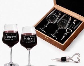 May include: A set of two clear wine glasses filled with red wine, each etched with the text Hubby or Wifey and EST 2025. The set includes a wooden box containing two additional glasses, a heart-shaped metal wine stopper, and a black corkscrew.
