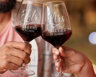 May include: Two clear glass wine glasses filled with red wine. One glass is etched with the text Mr. MILLERS 24.10.2024 and the other with Mrs. MILLERS 24.10.2024 in a white script font with decorative flourishes above the surname and date.