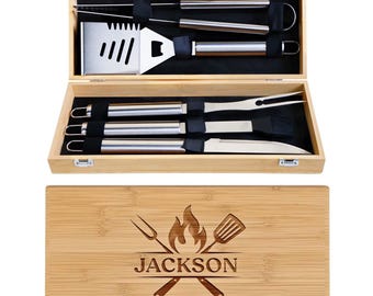 May include: A barbecue tool set in a light wood carrying case with metal latches. The case lid features a laser-engraved flame icon, crossed utensils, and the name JACKSON. Inside are stainless steel tongs, a spatula, a fork, a brush, and a knife.