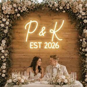 May include: A wedding reception scene with a couple seated at a table. The backdrop features a wooden wall with a neon sign displaying initials and the year "EST 2026". Floral arrangements and candles decorate the table and surrounding area.