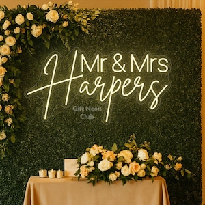Könnte beinhalten: Eine Hochzeitskulisse mit einem Neonschild mit der Aufschrift „Mr & Mrs Harpers“ vor einer grünen Heckenwand. Das Schild ist von weißen Rosen und Grün umgeben. Davor steht ein Tisch mit Kerzen und Blumen.