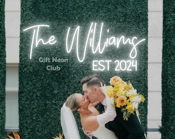 Könnte beinhalten: Ein Paar küsst sich vor einem Neonschild mit der Aufschrift "The Williams EST 2024" und darunter den Worten "Gift Neon Club". Das Paar steht vor einer grünen Wand mit gelben Blumen.