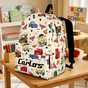 May include: A cream-coloured backpack featuring a repeating pattern of various cartoon cars, traffic cones, and checkered flags. The name Carlos is printed in bold black letters on the front pocket, accompanied by small black star shapes.
