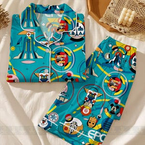 May include: A teal pajama set with a colorful cartoon design. The top has a collared neckline and white buttons. The pants and top feature a repeating pattern of cartoon characters, geometric shapes, and architectural elements.