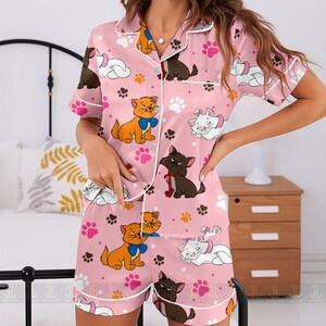 May include: Pink pajama set featuring a repeating pattern of cartoon cats, paw prints, and clouds. The short-sleeved top and shorts have white trim. The cats are in various colors, including orange, brown, and white.