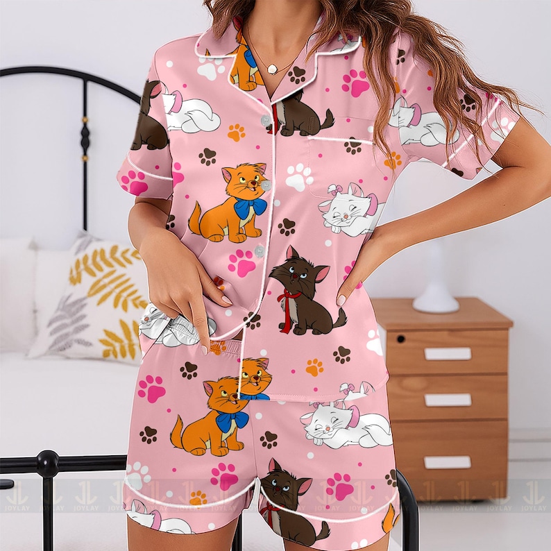 May include: Pink pajama set featuring a repeating pattern of cartoon cats, paw prints, and clouds. The short-sleeved top and shorts have white trim. The cats are in various colors, including orange, brown, and white.