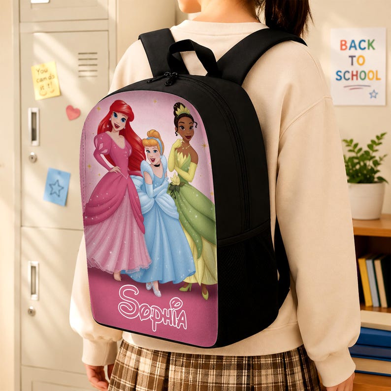 May include: A black backpack featuring a graphic print of three animated princesses in pink, blue, and green gowns on a pink background. The name Sophia is printed in white script at the bottom of the front panel. The bag has black shoulder straps.