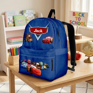 May include: A blue backpack with characters from the film Cars, and the name "Jack" on a red and white label. The backpack has black straps and a zip pocket. A "Back to School" sign is visible in the background.
