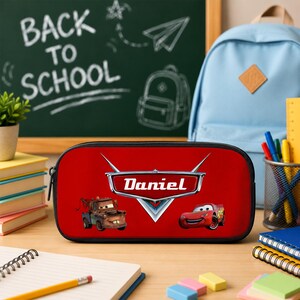 May include: A red rectangular pencil case featuring the Disney Pixar Cars logo with the name Daniel printed in white text. The case displays illustrations of the characters Mater and Lightning McQueen on a vibrant red background with a black zipper.