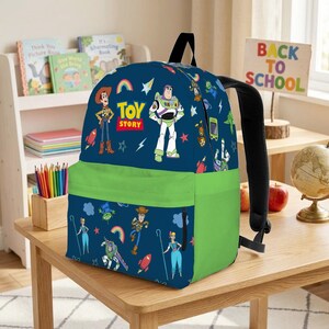 May include: A navy blue backpack with a green front pocket, featuring Toy Story characters. The backpack has black straps and the words "TOY STORY" are printed on the front. A sign that says "BACK TO SCHOOL" is in the background.
