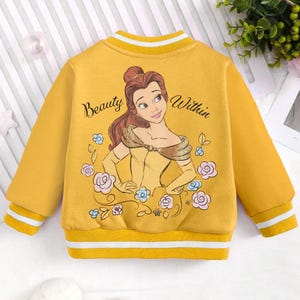 May include: Yellow bomber jacket with a cartoon image of Belle from Beauty and the Beast. The jacket has white stripes on the sleeves and collar. The text "Beauty Within" is printed on the jacket.