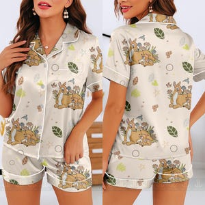 Op de afbeelding: Cr&egrave;mekleurige pyjamaset met een Bambi en Stampertje print. De top met korte mouwen en de shorts hebben witte biezen en knoopsluitingen. De set bevat een herhalend patroon van herten, bloemen, bladeren en vogels.