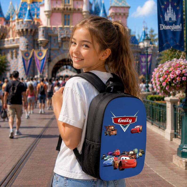 K&ouml;nnte beinhalten: Ein schwarzer Rucksack mit einer blauen Frontpartie, die eine Grafik verschiedener animierter Autos und den Namen Emily in einem rot-wei&szlig;en Logo zeigt. Der Rucksack verf&uuml;gt &uuml;ber schwarze Schultergurte und eine seitliche Netztasche zur Aufbewahrung.