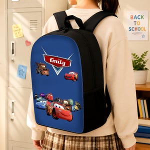 May include: A black backpack featuring a blue front panel with a graphic print of various animated car characters. A red emblem at the top displays the name Emily in white text. The bag includes black shoulder straps and side mesh pockets.